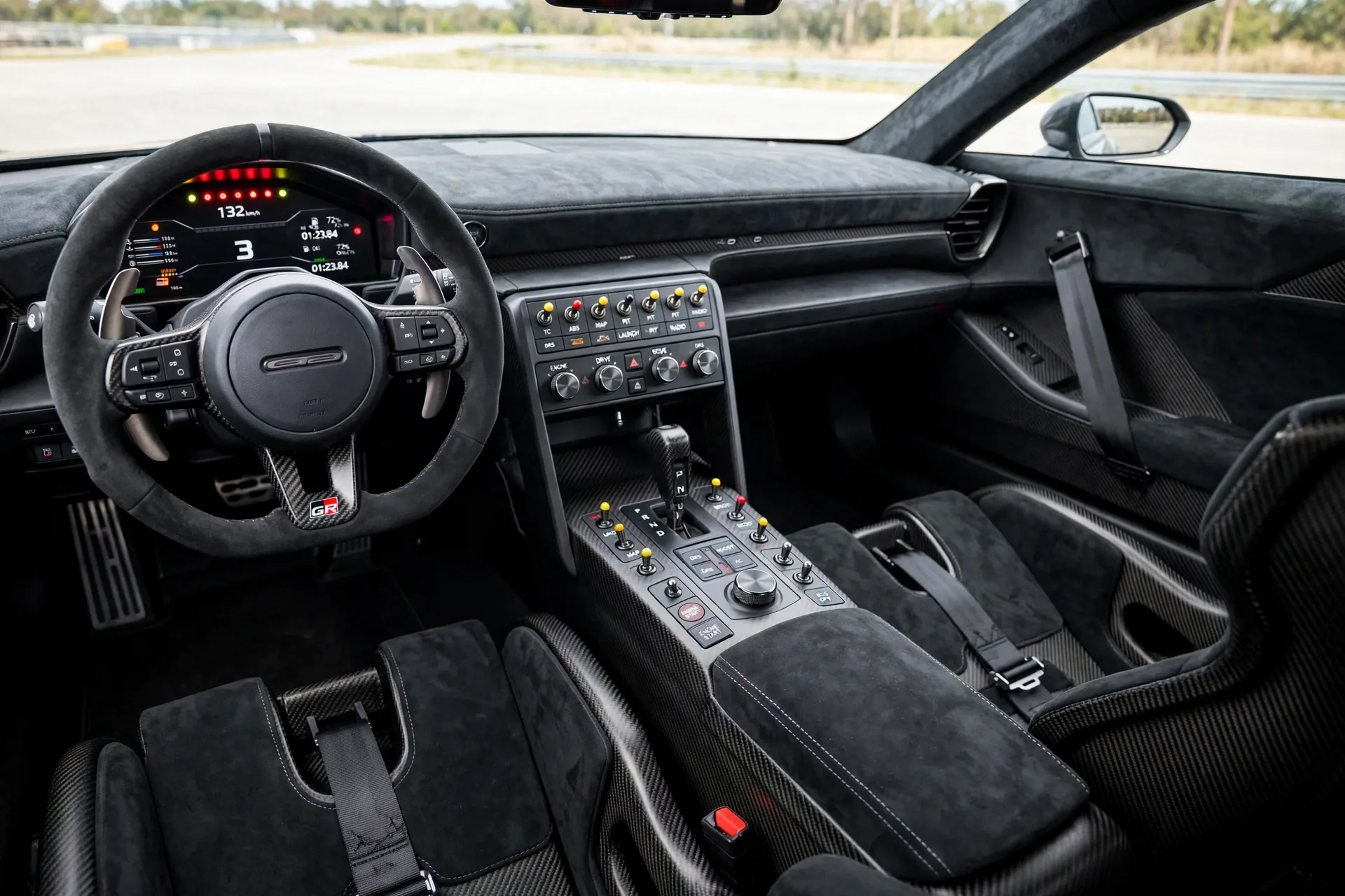 Driver-focused Toyota GR GT cockpit with carbon bucket seats, a shift-light digital cluster, a flat-bottom steering wheel, and a console full of physical toggle switches